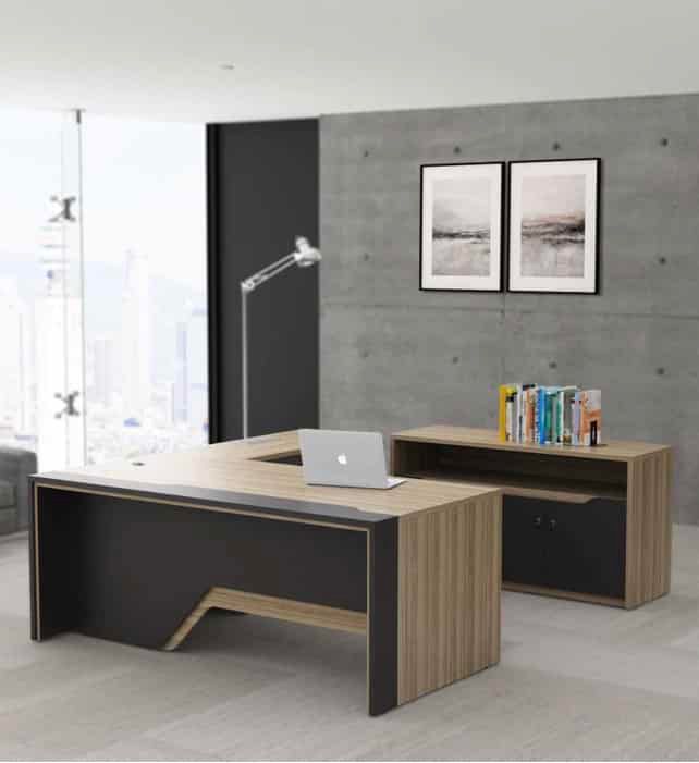 "A modern L-shaped executive desk in light teak wood with an asymmetrical dark grey front modesty panel and integrated side return."