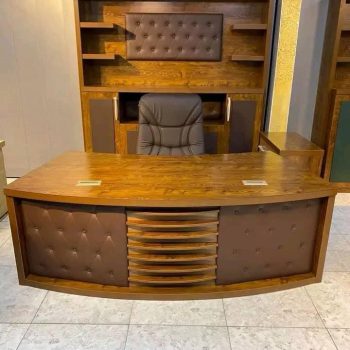 "A curved wooden executive desk in a medium oak finish, featuring brown button-tufted leather panels and a central slatted design on the front modesty panel."