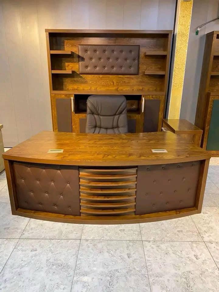 "A curved wooden executive desk in a medium oak finish, featuring brown button-tufted leather panels and a central slatted design on the front modesty panel."