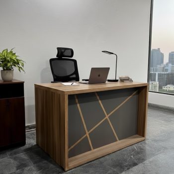 "A contemporary rectangular staff desk in a warm light oak finish, featuring a thick boxed frame and a charcoal grey front panel with intersecting geometric wood line details."