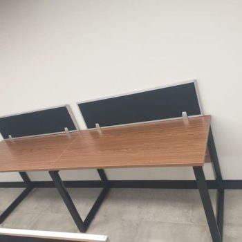 "A linear 2-person side-by-side office workstation desk featuring warm teak wood-grain tabletops, an architectural black metal trapezoidal loop frame, and rear-mounted black privacy divider screens with silver aluminum borders."