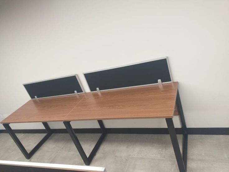 "A linear 2-person side-by-side office workstation desk featuring warm teak wood-grain tabletops, an architectural black metal trapezoidal loop frame, and rear-mounted black privacy divider screens with silver aluminum borders."