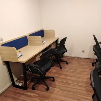 "A row of linear modular office cubicle desks featuring a light wood-grain laminate finish, integrated curved side privacy panels with blue fabric backboards, black metal legs, and built-in under-desk drawer units."