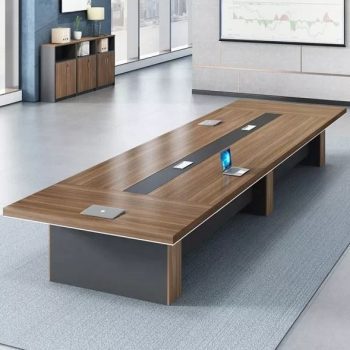 "A long, rectangular executive boardroom desk featuring a warm walnut wood-grain top with a recessed black center media track, supported by three thick charcoal-grey pedestal bases."